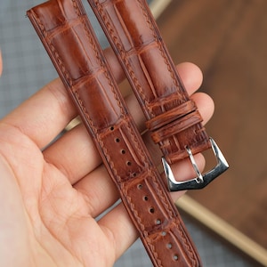 May include: Brown leather watch straps with a crocodile pattern. The straps have a silver-colored buckle and are held in a hand. The leather has a textured appearance and is stitched along the edges.