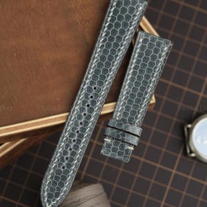 Peut inclure: Un bracelet de montre gris foncé présentant un motif hexagonal texturé et des coutures claires. Le bracelet se compose de deux pièces, l'une avec plusieurs trous de réglage et l'autre avec deux passants coulissants, pour une montre.