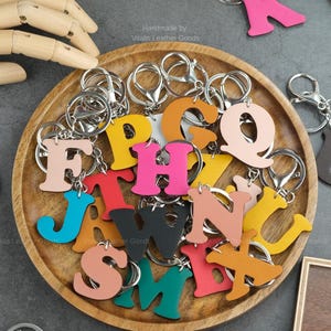 Könnte beinhalten: Eine Sammlung bunter Leder-Buchstaben-Schlüsselanhänger in verschiedenen Farbtönen, darunter Pink, Gelb und Blau, wird auf einem Holztablett präsentiert. Jeder Buchstabe ist an einem silbernen Schlüsselring befestigt. Der Text "Handmade by Vitalis Leather Goods" ist sichtbar.