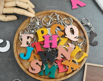May include: A collection of colorful leather letter keychains in various hues, including pink, yellow, and blue, are displayed on a wooden tray. Each letter is attached to a silver keyring. The text "Handmade by Vitalis Leather Goods" is visible.