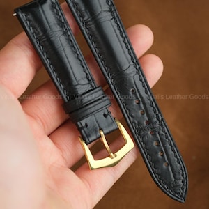 Puede incluir: Correas de reloj de cuero negro con un patrón texturizado y hebilla dorada. Las correas están cosidas con hilo en contraste. El texto "Handmade by Vitalis Leather Goods" es visible en la parte superior de la imagen.