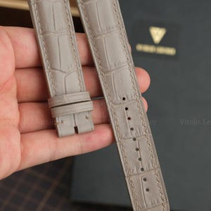 Può includere: Un cinturino per orologio grigio chiaro con una trama testurizzata in pelle di alligatore e cuciture abbinate. Il cinturino è composto da due pezzi, uno con un'estremità a fibbia con due passanti e l'altro con una serie di fori di regolazione.