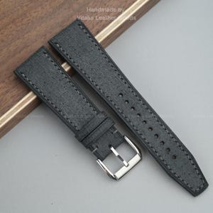 Pode incluir: Um par de pulseiras de relógio cinza escuro com costuras cinza claro. As pulseiras têm uma aparência texturizada e uma fivela prateada. O texto "Handmade by Vitalis Leather Goods" é visível no topo.