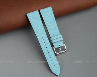 May include: Light blue leather watch straps with a silver buckle. The straps are made of textured leather and have multiple holes for adjustment. The background is a mix of brown and gray.