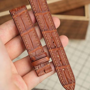 Könnte beinhalten: Ein braunes Leder-Uhrenarmband mit einer geprägten Alligator-Struktur. Das Armband verfügt über helle Kontrastnähte an den Rändern, eine feste Lederschlaufe und mehrere vorgestanzte Löcher zur individuellen Größenanpassung.