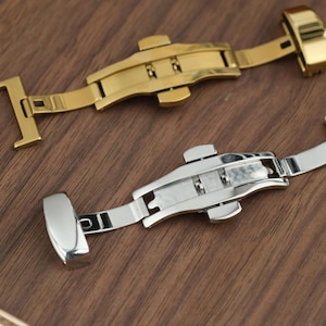 May include: Two watch clasps, one gold-toned and one silver-toned, are displayed on a wooden surface. The clasps are open, showcasing their intricate design and locking mechanisms. The gold clasp is positioned above the silver clasp.