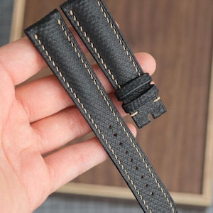 Puede incluir: Correa de reloj de cuero negro con costuras beige. La correa tiene una superficie texturizada y una serie de agujeros para ajustar. La correa del reloj está sujeta en la mano de una persona.