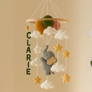 以下が含まれることがあります： 木製のリング、フェルトの風船、雲、星が付いたベビーモバイル。ベージュの耳と鼻を持つ灰色の象が下に吊り下げられています。「CLARIE」という名前は緑色の文字で書かれています。 赤ちゃんの部屋用。