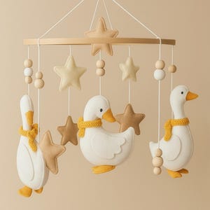Pode incluir: Móbile de bebê com três gansos brancos com bicos, pés e cachecóis amarelos. Estrelas bege e contas de madeira suspensas em um aro de madeira. Decoração para o quarto do bebê.