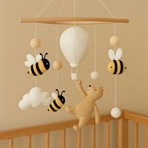 May include: A baby mobile featuring a wooden frame with hanging felt decorations. The mobile includes a white hot air balloon, a teddy bear, bees with black and yellow stripes, white clouds, and wooden spheres. The mobile is suspended above a wooden crib.