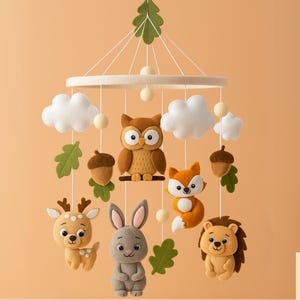 Op de afbeelding: Een handgemaakte baby mobile met bosdieren. De mobiel bevat een uil, vos, hert, konijn en egel, samen met witte wolken, eikels en groene bladeren. Opgehangen aan een witte ring.