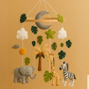 May include: A handmade baby mobile featuring felt safari animals, including an elephant, giraffe, and zebra, along with a felt moon, clouds, and leaves. The mobile is suspended from a wooden hoop with a neutral color palette.