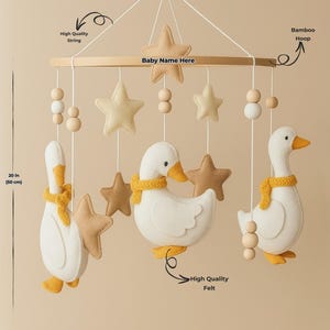 Op de afbeelding: Een baby mobile met drie witte vilten ganzen met gele accenten en stervormige ornamenten. De mobiel is opgehangen aan een houten frame en meet 50 cm in lengte. Inclusief hoogwaardig touw en vilt.
