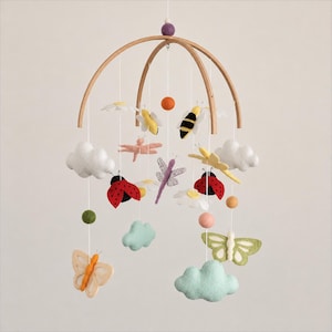 May include: A hanging mobile featuring a wooden arched frame with felted wool figures including white clouds, red ladybugs, yellow bees, a pink dragonfly, a purple dragonfly, a green butterfly, a peach butterfly, and white daisies.