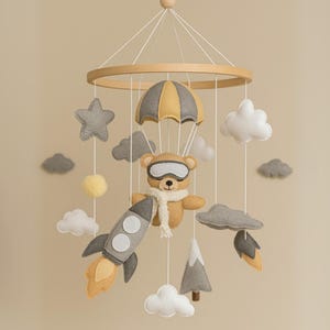 Op de afbeelding: Een baby mobile met een teddybeer met een bril en sjaal, opgehangen aan een houten ring. De mobile bevat een raket, wolken, sterren, een parachute en een berg, allemaal in zachte, neutrale kleuren.