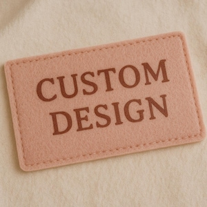 May include: A rectangular, light pink felt patch with the words "CUSTOM DESIGN" printed in brown. The patch has a stitched border and is set against a cream-colored fabric background.