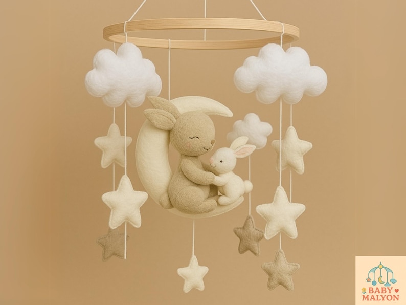 May include: A nursery mobile with a wooden hoop, felt clouds, stars, and a moon. Two plush bunnies are seated on the moon. The mobile is in neutral colours, suitable for a baby's room.