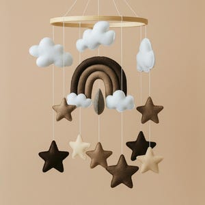 May include: A baby mobile featuring a wooden ring with hanging felt clouds, stars, and a rainbow in shades of brown and white. The stars are in various shades of brown and cream. The mobile is suspended against a neutral background.
