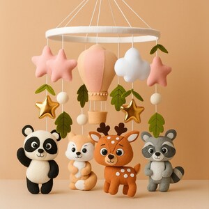 May include: A baby mobile featuring felt animal figures including a panda, fox, deer, and raccoon. The mobile also includes a hot air balloon, stars, and clouds, all suspended from a white ring. The color palette is soft, with pastel pinks, golds, and greens.