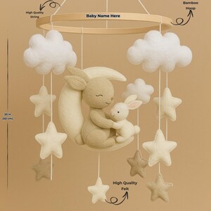 May include: A baby mobile with a beige bunny and a smaller white bunny embracing on a crescent moon. The mobile includes white clouds and star shapes, suspended from a wooden hoop. The mobile is 50 cm in length.