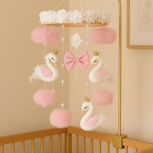 Op de afbeelding: Een baby mobile met witte zwanen met gouden kronen, roze pompons en een roze strik. De mobile hangt aan een goudkleurige arm en is ontworpen om boven een wieg te hangen.