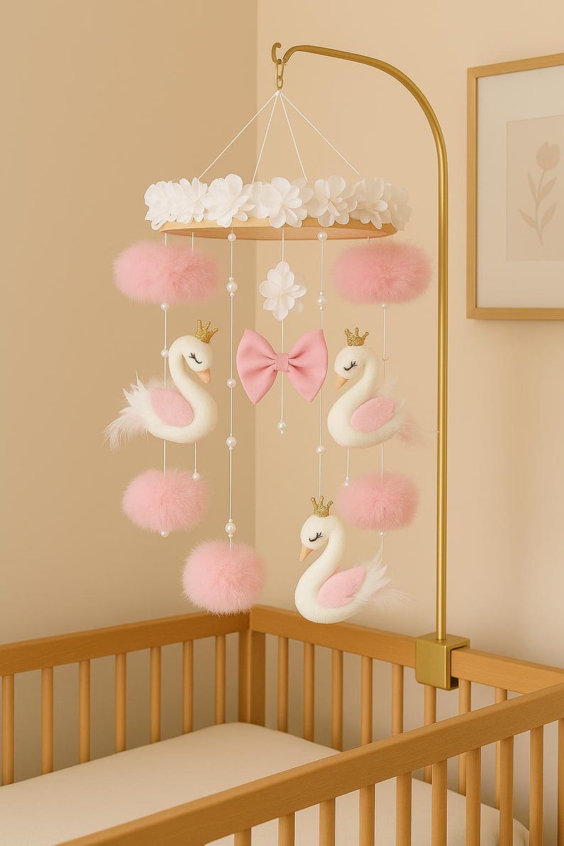 Op de afbeelding: Een baby mobile met witte zwanen met gouden kronen, roze pompons en een roze strik. De mobile hangt aan een goudkleurige arm en is ontworpen om boven een wieg te hangen.