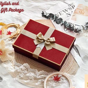 May include: A red gift box with a gold ribbon and bow, presented on a woven mat. The image includes decorative elements like a gold frame, a starfish, and lace trim, suggesting a gift package.