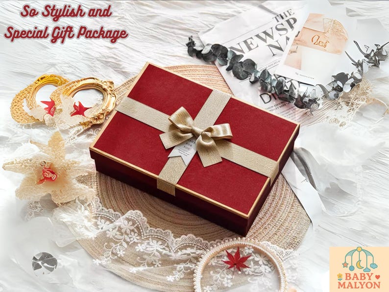 May include: A red gift box with a gold ribbon and bow, presented on a woven mat. The image includes decorative elements like a gold frame, a starfish, and lace trim, suggesting a gift package.