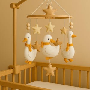 Op de afbeelding: Een houten baby mobile met drie witte eendenfiguren met gele sjaals, afgewisseld met bruinachtige sterren. De mobiel hangt boven een houten wieg met een witte matras. De eenden hebben gele snavels en voeten.