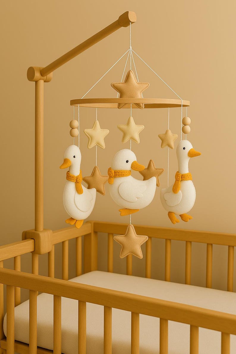Op de afbeelding: Een houten baby mobile met drie witte eendenfiguren met gele sjaals, afgewisseld met bruinachtige sterren. De mobiel hangt boven een houten wieg met een witte matras. De eenden hebben gele snavels en voeten.