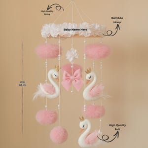 Op de afbeelding: Een baby mobile met witte zwanen met gouden kronen, roze accenten en een roze strik. De mobile bevat roze pompons en is opgehangen aan een bamboe hoepel. De mobile is 50 cm lang.