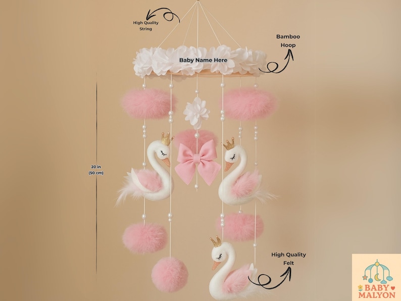 Op de afbeelding: Een baby mobile met witte zwanen met gouden kronen, roze accenten en een roze strik. De mobile bevat roze pompons en is opgehangen aan een bamboe hoepel. De mobile is 50 cm lang.
