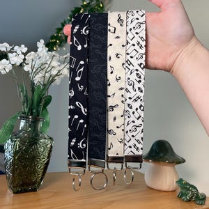 May include: Four fabric keychains with musical note patterns in black and white are displayed. Each keychain has a metal clasp and a key ring. The patterns include treble clefs, eighth notes, and other musical symbols. The keychains are held up by a hand.
