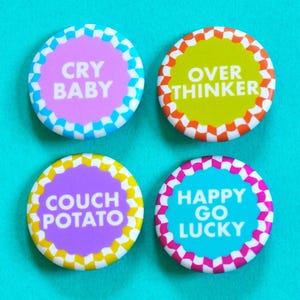 Könnte beinhalten: Vier bunte runde Knöpfe mit weißen karierten Rändern und weißem Text. Die Knöpfe zeigen "Cry Baby", "Over Thinker", "Couch Potato" und "Happy Go Lucky".