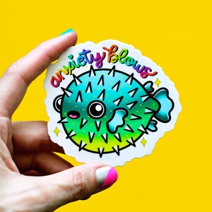 May include: A white sticker with a colorful cartoon pufferfish and the text "anxiety blows". The pufferfish is green, blue, and yellow with black eyes and a white mouth.