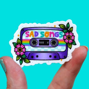 May include: A purple cassette tape sticker featuring the text SAD SONGS in colorful letters. The design includes a rainbow stripe, a light blue checkered pattern, and pink flowers with green leaves on the left and right sides of the tape.