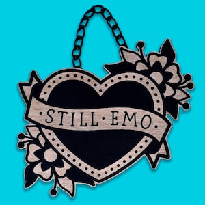 May include: A wooden heart-shaped sign with a black heart and floral details. The sign has a chain for hanging and the text "STILL EMO" on a banner.