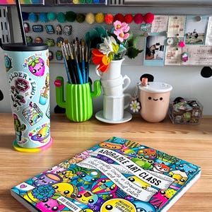 Pode incluir: Um espa&ccedil;o de trabalho com materiais de arte coloridos, incluindo um copo decorado, um porta-pinc&eacute;is em forma de cacto e um livro intitulado "Adorable Art Class". A capa do livro &eacute; preenchida com ilustra&ccedil;&otilde;es de desenhos animados.
