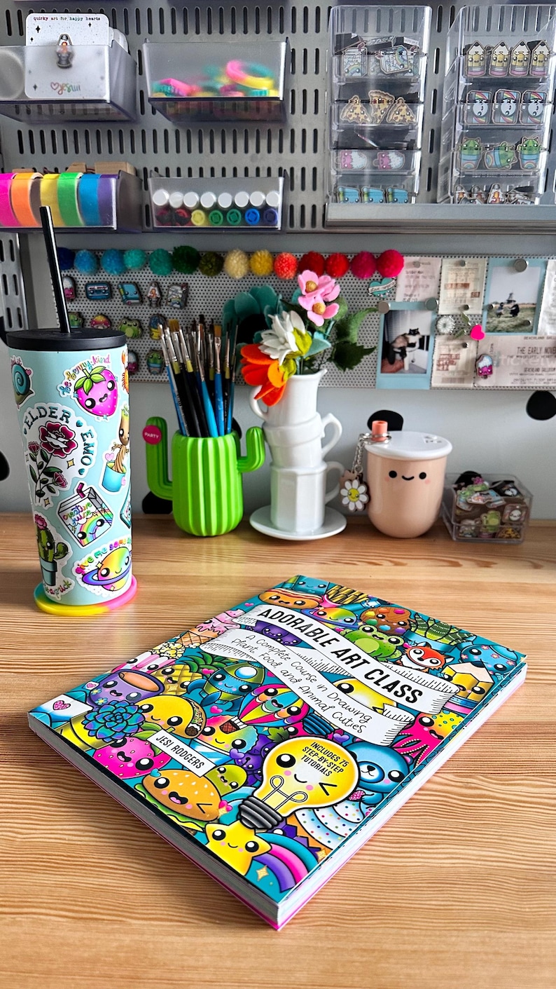 Pode incluir: Um espa&ccedil;o de trabalho com materiais de arte coloridos, incluindo um copo decorado, um porta-pinc&eacute;is em forma de cacto e um livro intitulado "Adorable Art Class". A capa do livro &eacute; preenchida com ilustra&ccedil;&otilde;es de desenhos animados.
