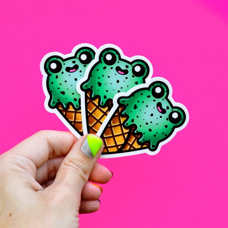 Pode incluir: Tr&ecirc;s adesivos de cone de sorvete de sapo verde. Cada adesivo apresenta um sapo com um rosto sorridente e um cone de sorvete verde com um cone de waffle.