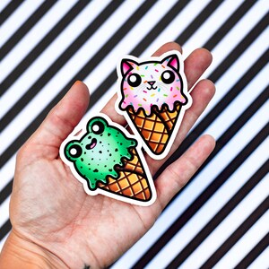 Pode incluir: Dois adesivos de cone de sorvete, um com um sapo verde e o outro com um gato rosa. Ambos os adesivos t&ecirc;m granulado no topo e est&atilde;o sobre um fundo listrado preto e branco.