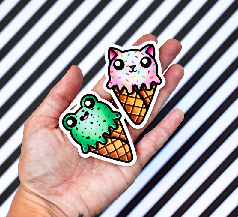 Pode incluir: Dois adesivos de cone de sorvete, um com um sapo verde e o outro com um gato rosa. Ambos os adesivos t&ecirc;m granulado no topo e est&atilde;o sobre um fundo listrado preto e branco.