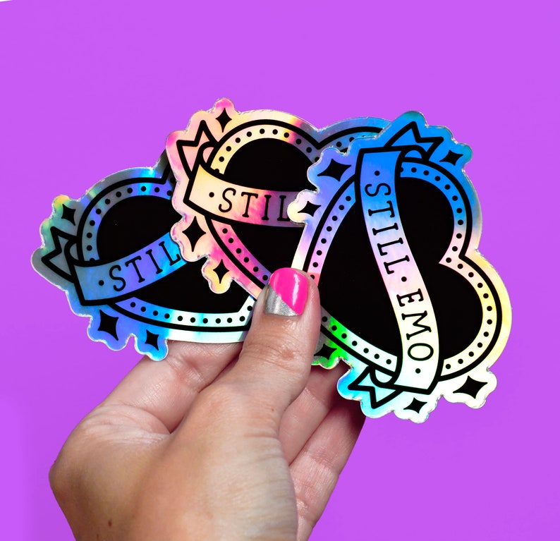 May include: Holographic heart-shaped stickers with black centres and the words "STILL EMO" on a banner. The stickers have a black border with star and dot details. The background is a solid purple.