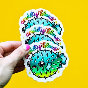 May include: Three colorful stickers with a cartoon pufferfish and the text "anxiety blows" in rainbow letters. The stickers are layered on top of each other.