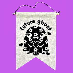 Könnte beinhalten: Ein graues Filzbanner mit den schwarzen Worten "future ghost". Das Banner zeigt einen schwarzen Geistercharakter mit weißen Blumendetails, der ein Herz hält. Das Banner wird von einem Holzstab und einer lila-weißen Schnur gehalten.