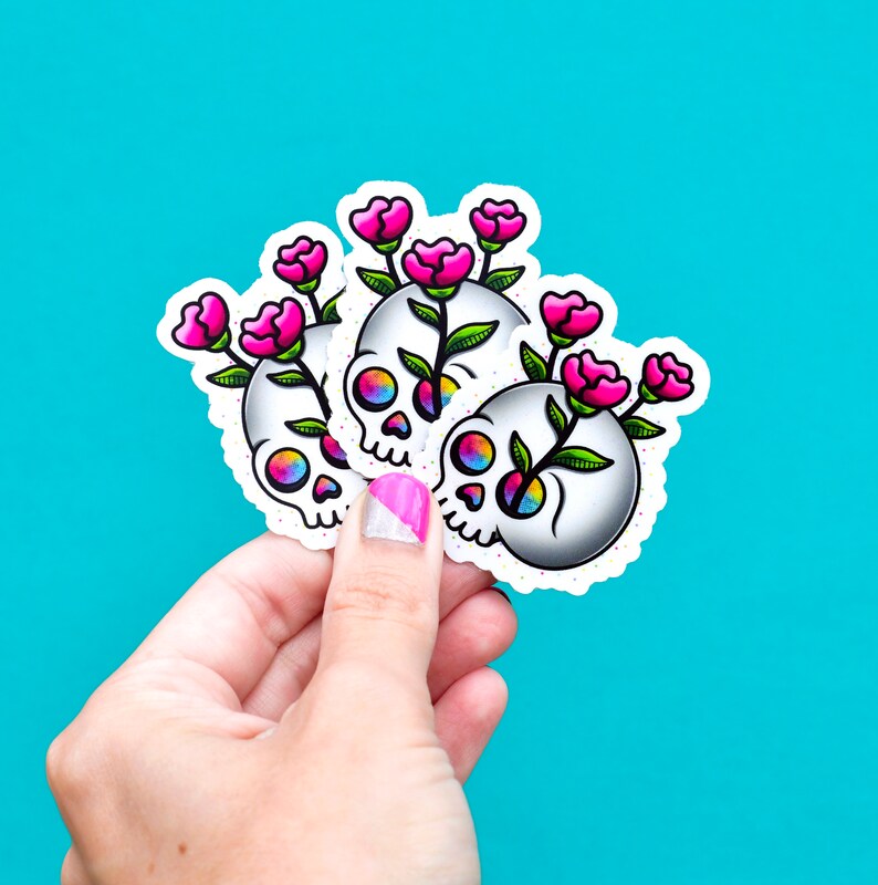 May include: Three colorful skull stickers with pink flowers growing out of the top of each skull. The stickers are on a blue background.