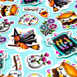 May include: A collection of colorful Halloween-themed stickers on a light blue background. The stickers feature various designs, including a witch on a broom, skulls with flowers, and a television with the text "snacks & spooky movies".