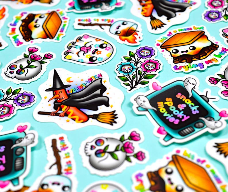 May include: A collection of colorful Halloween-themed stickers on a light blue background. The stickers feature various designs, including a witch on a broom, skulls with flowers, and a television with the text "snacks & spooky movies".