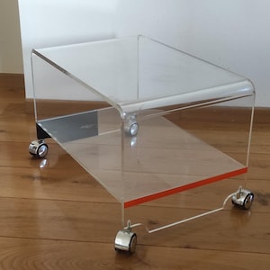 May include: A clear acrylic side table with a rectangular top and a lower shelf. The table has a red accent on the bottom edge and sits on four chrome caster wheels. The table is designed with a modern aesthetic.