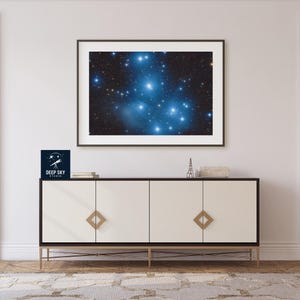 May include: A framed photograph of a star cluster in a dark blue sky. The image is displayed on a white wall above a white cabinet with gold accents. The cabinet has three doors and a black base. A small logo with the text "DEEP SKY STUDIO" is visible on the left side of the image.